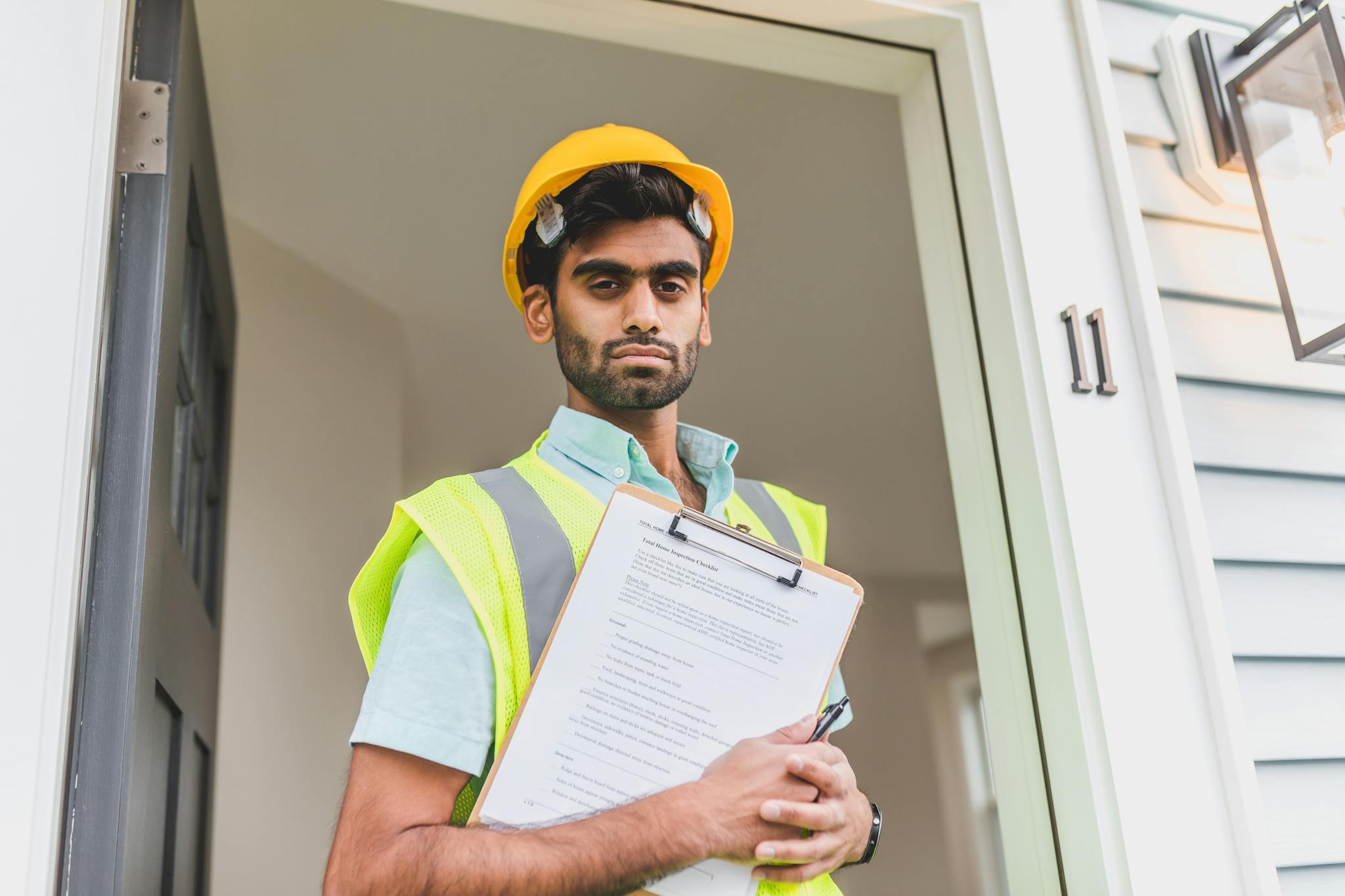 South Asian construction worker at a doorway, holding a clipboard, wearing a hardhat and safety vest.